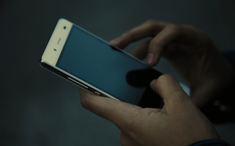 A person's hands interacting with a smartphone in a dimly lit environment, suggesting the use of mobile devices for various purposes in different settings.